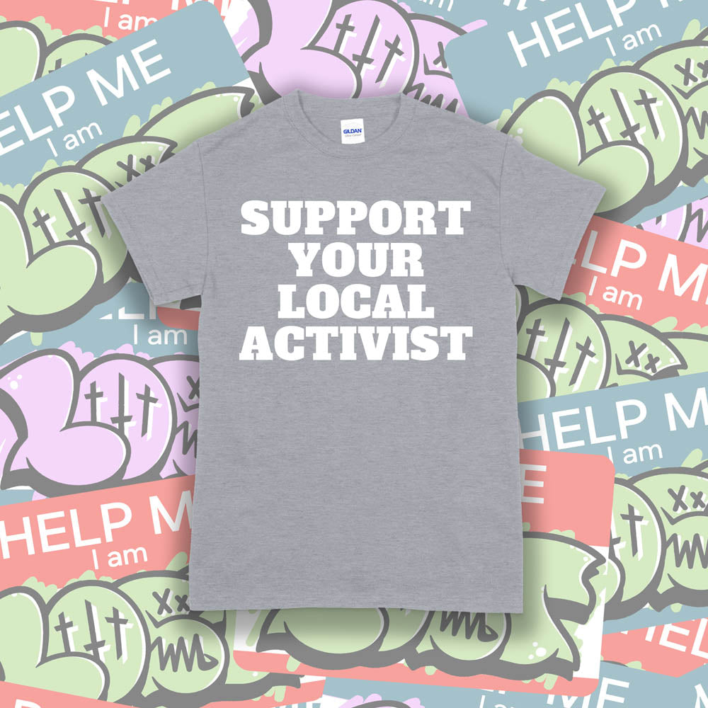 The original Support Your Local Activist t-shirt. Bold white letters screen printed on a gray shirt. The shirt is displayed against a wall of colorful graffiti stickers.