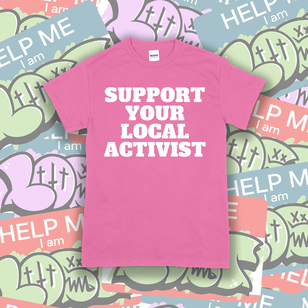The original Support Your Local Activist t-shirt. Bold white letters screen printed on a pink shirt. The shirt is displayed against a wall of colorful graffiti stickers.