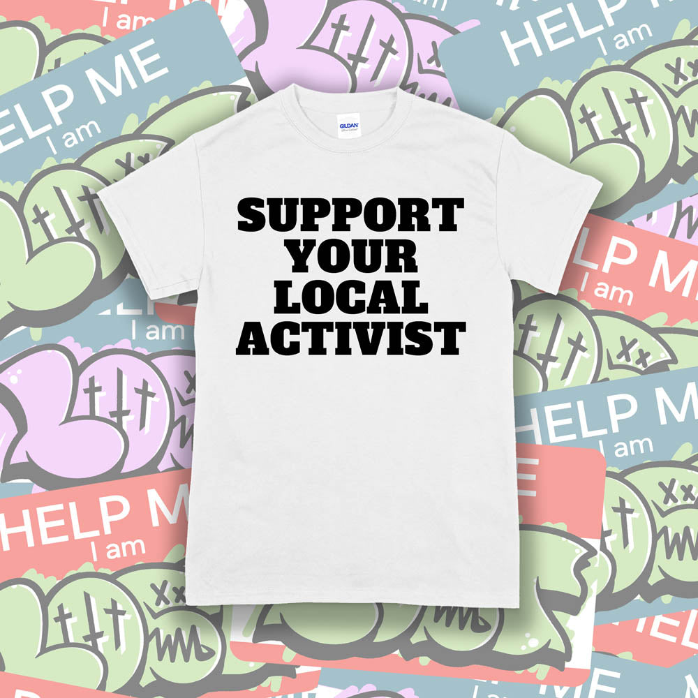 The original Support Your Local Activist t-shirt. Bold black letters screen printed on a white shirt. The shirt is displayed against a wall of colorful graffiti stickers.